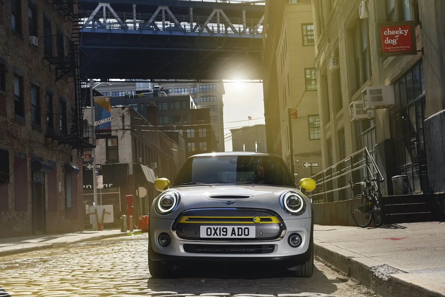 A MINI Electric on a cobbled city street beneath a railway bridge in low sun
