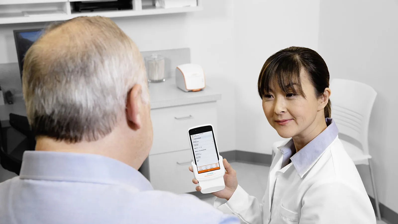 A patient holding the handheld Atellica DCA analyzer in an exam room while a clinician sits opposite